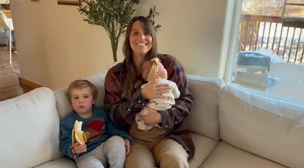 A toddler kisses his newborn sister while his mother captures the moment on video