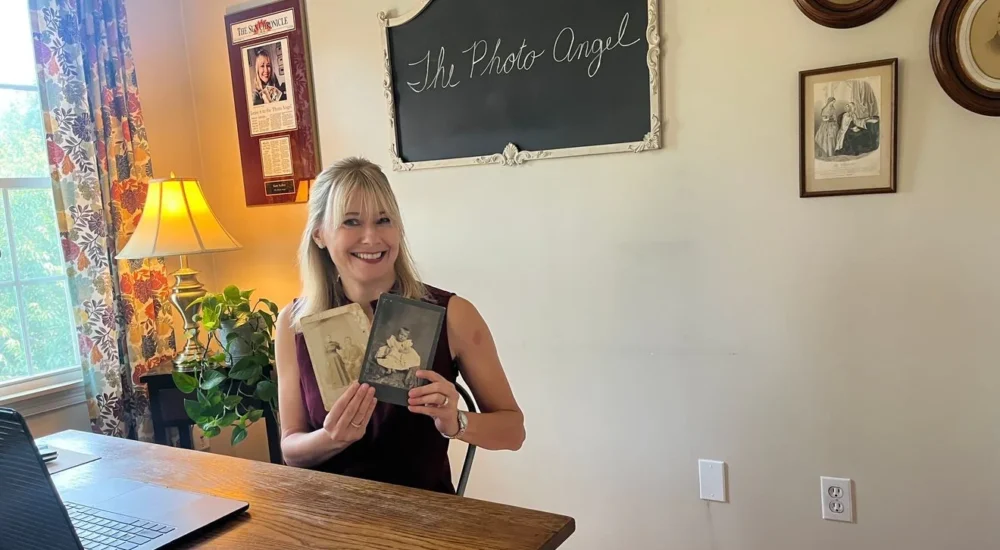 Woman holding a vintage family photo while standing beside a map marked with pins across the United States